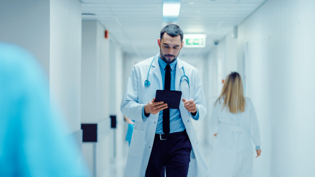 Doctor walking through a hospital corridor while reviewing information on a tablet.