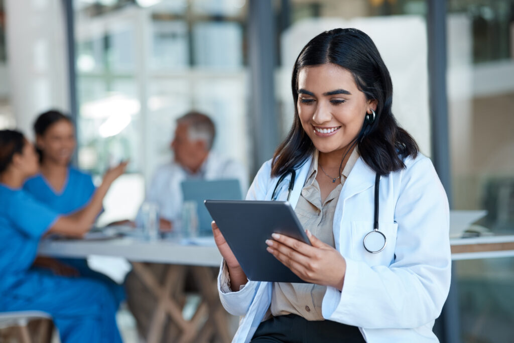 Medical professional holding a tablet in a clinic, with people working at computers in the background.
